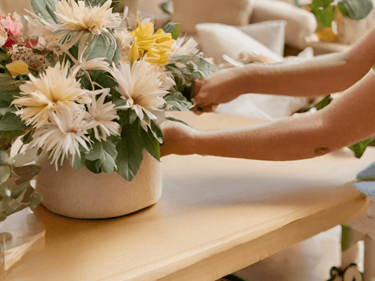 A person is arranging flowers in a vase on a table. The flowers are mostly chrysanthemums, with a few other types of flowers mixed in. The person is carefully placing each flower in the vase, and the arrangement is starting to look beautiful.