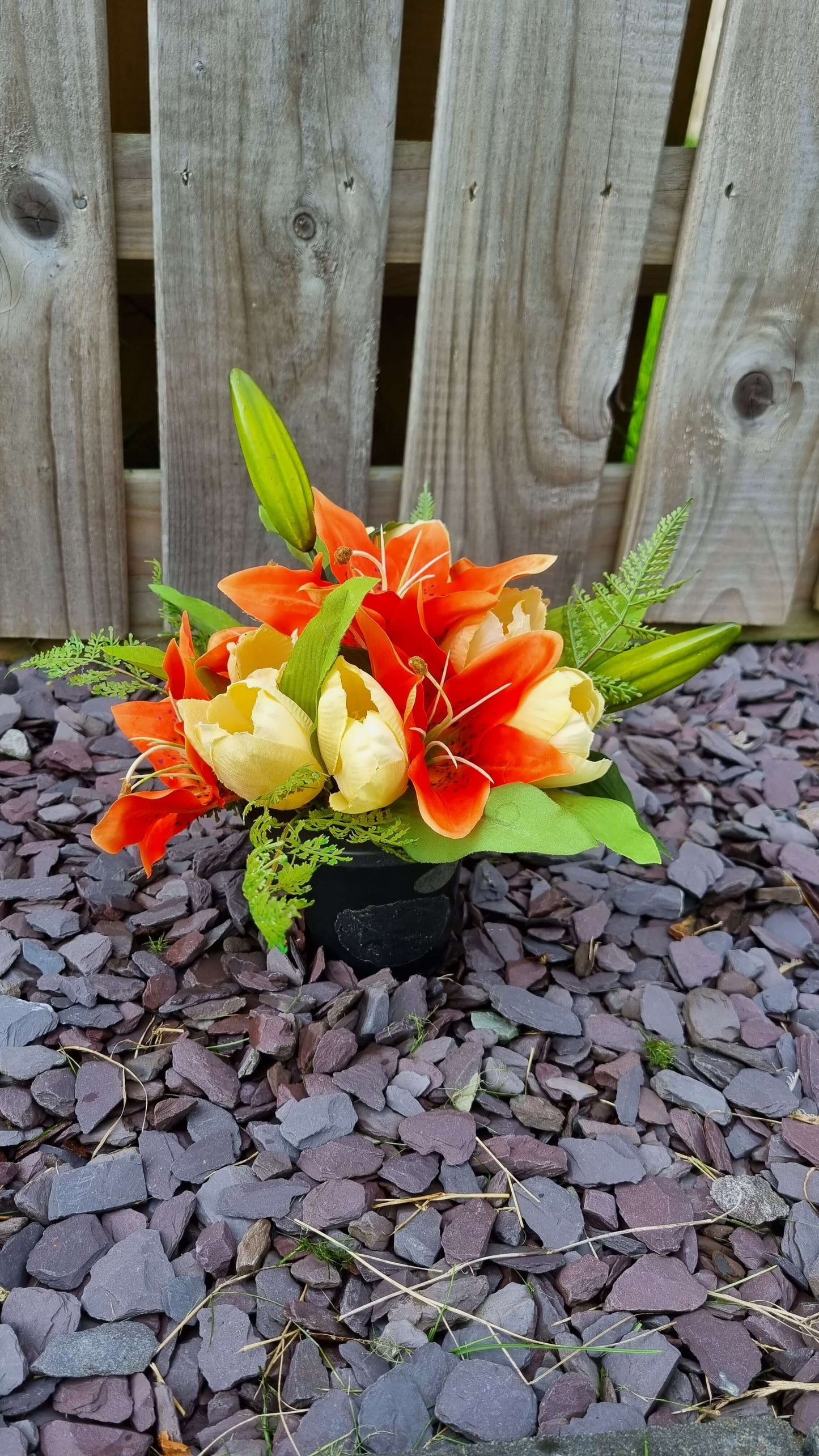 A photo of an orange lily memorial grave pot sitting on top of small purple and grey stones. The grave pot is filled with artificial orange lilies and foliage.