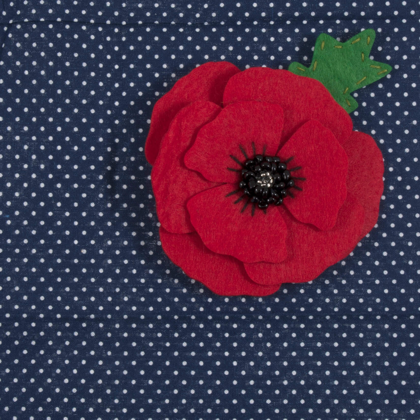 The image shows a red felt poppy on a blue polka dot background.