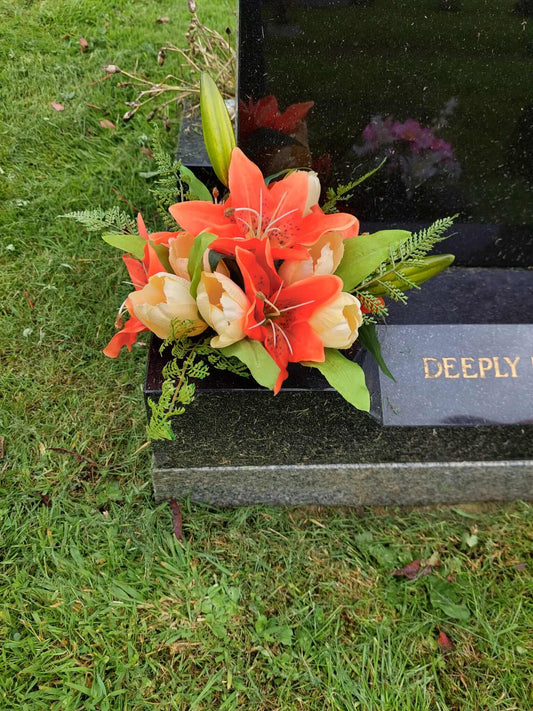 A close-up photo of a bunch of orange lilies sitting on the side of a grave. The lilies are in full bloom and their bright orange color contrasts with the green foliage and the black headstone.