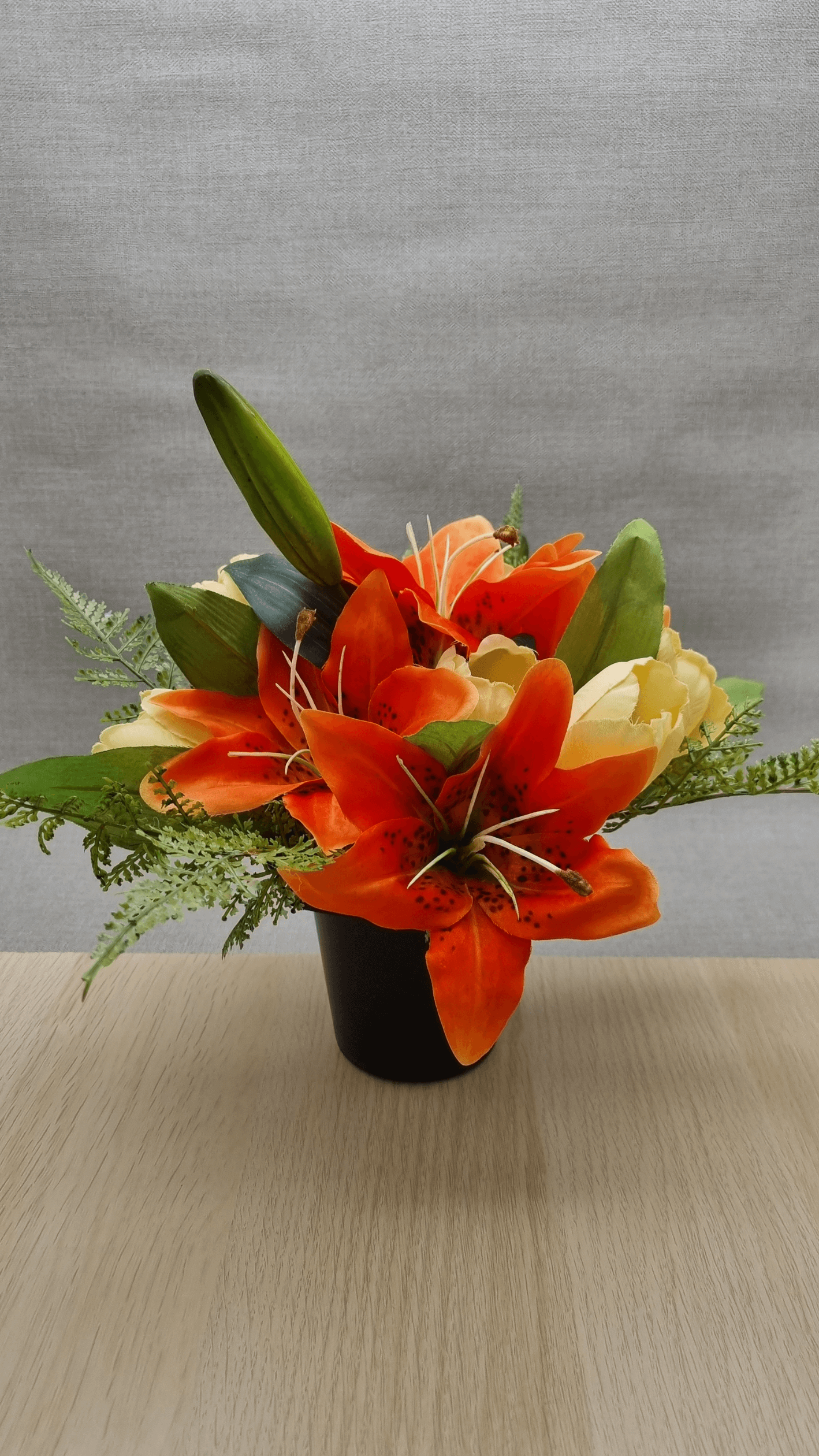 A bouquet of orange lilies in a black grave pot on a wooden table. A light green background nestles behind. 